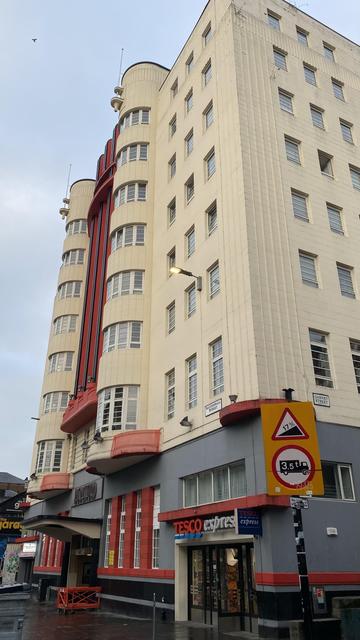 An art deco block of flats with cream harling and a big red stripe down the centre of the building which ends in an awning. There’s a road sign and some street furniture in the foreground.