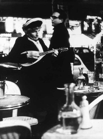 The image is a black and white photograph. In the center of the image, there is a person who appears to be a man playing an instrument. He is dressed in clothing that suggests a style from an earlier era, possibly mid-20th century, and he has his eyes closed, which might indicate he is performing or deeply engaged in the music. The setting is a bar or restaurant with tables and chairs visible in the background, suggesting a public dining area. There is also a bottle on one of the tables, further indicating that this is a social setting.

There are two other people present in the image; one appears to be standing near a table, possibly interacting with it, while the other person is sitting at a bar counter. The individual playing the instrument is slightly off-center and occupies a larger portion of the frame than the other people. The focus on this central figure suggests that he is the main subject of the photograph.

The image has a grainy texture which is characteristic of black and white photography, especially when it comes to capturing older scenes or vintage aesthetics. The overall composition of the image places the musician as the focal point against a background that includes other patrons in the establishment.