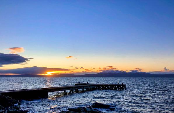 A scenic view of a pier extending into the water at sunset, with colorful skies and mountains in the background. Orange and blue hues dominate the scene, reflecting on the water's surface.