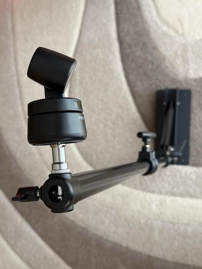 The image features a webcam mounted on a black mounting arm, showcasing a swivel joint and adjustable components. The background consists of a softly textured surface.