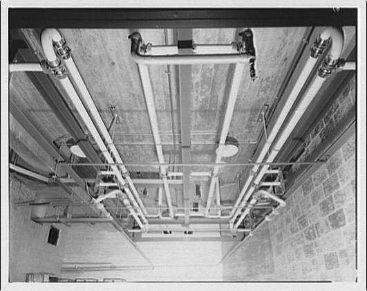 The image is a collage of two photographs taken from different angles, showcasing an industrial setting with pipes and valves.
In the upper photograph, we see an overhead view of the room where multiple metal pipes are running along the ceiling. These pipes have various valves attached to them at intervals. The pipe arrangement suggests a complex system or network, possibly for transporting water or other fluids.
The lower photograph offers a ground-level perspective, allowing us to see more details of the room's structure. It shows a concrete floor supporting the weight of the pipes and valves above. The layout of the piping suggests it might be part of an industrial setup, possibly for manufacturing or processing purposes.
Both photos are in black and white, which gives them a slightly nostalgic feel. There is no visible text within the image to provide additional context or information.