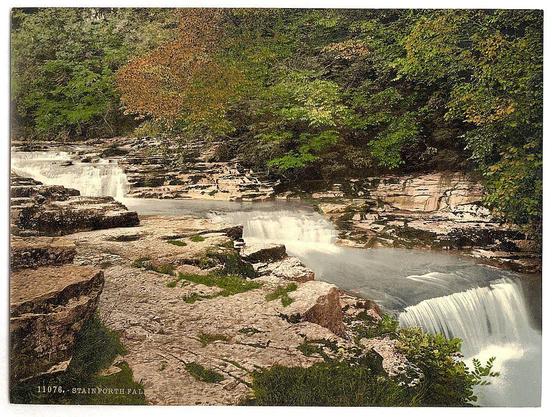 The image presents a captivating scene of a waterfall cascading down a rocky cliff. The fall, located on the right side of the photo, is surrounded by lush greenery and towering trees that add depth to the landscape. A river meanders through the bottom left corner of the frame, creating a serene atmosphere. The waterfall's position relative to the river suggests it may be providing the river with its source of water. On the right side of the photo, a small bridge can be seen, possibly serving as a connection between two parts of the landscape or as an access point for visitors to the natural wonder. The colors in the image are predominantly green and brown, indicating the presence of abundant vegetation and rocky terrain. There is no visible text within the image. The overall composition of the image suggests it was taken during daylight hours.