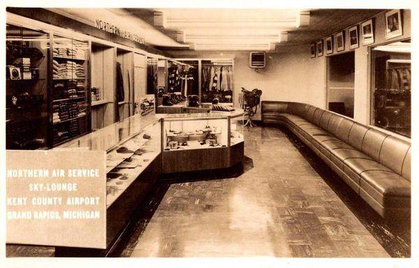 he final postcard shows the interior of the Northern Air Service Sky-Lounge located inside the terminal. The image captures display cases filled with travel goods, clothing, and accessories, as well as a long curved seating area. Framed aviation photographs line the wall above. This retail and lounge space reflects mid-20th-century airport amenities, which combined passenger services with local concessions to create a comfortable preflight environment.