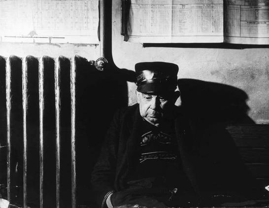 The image shows a black and white photograph of an older man sitting alone on a bench. He appears to be wearing a dark jacket, possibly with a collar, and a cap. His posture suggests he might be deep in thought or waiting for someone. Behind him, there's a wall with what looks like a radiator and some papers or documents pinned up. The man is the central figure in the image, and his position on the bench gives him prominence. There are no other people visible in the photo. The setting seems to be indoors, possibly a public space such as a waiting area or a station.