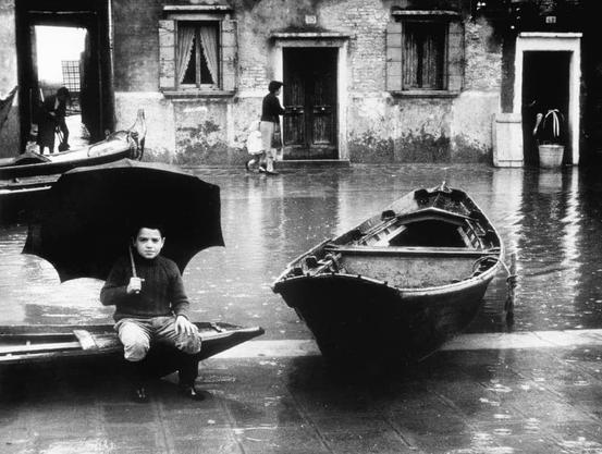 The image depicts a scene of a person sitting on a bench next to a body of water, which appears to be a canal or river. The individual is holding an umbrella and seems to be waiting or resting. There are several boats visible in the water, suggesting that this might be a common mode of transportation or a popular spot for boating activities. In the background, there are buildings with a rustic appearance, possibly indicating an older city or town. The ground is wet, which could imply recent rainfall or flooding. The overall atmosphere of the image suggests a calm and serene setting.