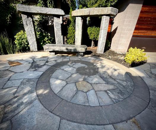 Apaved area or patio with a spiral motif. At the edge is a long low stone bench and behind it two stone trilithons of narrow stones.