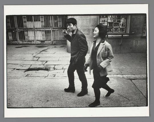 The image is a black-and-white photograph depicting two individuals walking side by side on a paved street. The person on the left is smoking a cigarette and is dressed in a dark jacket and pants, paired with dark shoes. The person on the right is wearing a light-colored coat over a dark outfit, with dark shoes, and is walking with a slight smile. They appear to be holding hands as they walk. The background includes a wall with a window, which has a grid-like pattern and what seems to be some posters or advertisements displayed on it. The ground is made up of large, uneven paving stones, and there is a visible crack or line on the pavement. The overall atmosphere of the image suggests a candid street scene, possibly from a past era given the style of clothing and the black-and-white format of the photograph.