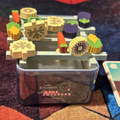 The bottom of the Barbecubes Veggie tin box with the wooden grill pieces on top. On the grill pixelated wooden vegetables stacked haphazardly.