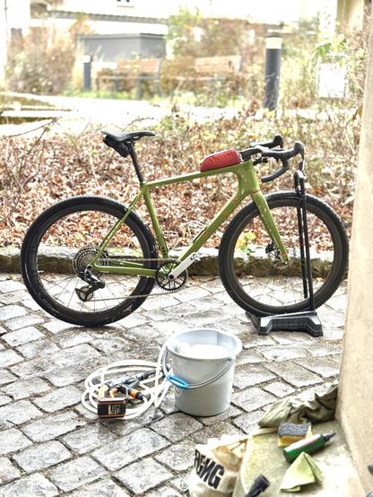 A green bicycle is parked on cobblestone ground, accompanied by a bucket, cleaning tools, and a hose. The background includes dried leaves and distant benches, suggesting an outdoor setting.