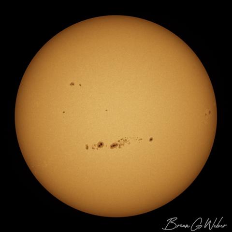 A full disk monochrome image of the sun, stacked from 750 frames of video and colorized orange. The main feature is a tight band of sunspots right in the middle just below the equator.