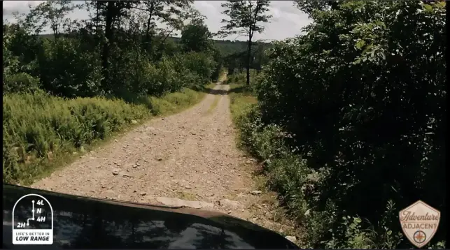 View over the hood of the FJ Cruiser climbing a rocky dirt road on Berry Mountain, surrounded by summer-green trees, with a ‘Life’s Better in Low Range’ graphic and Adventure Adjacent logo in the bottom left and right corners, respectively.