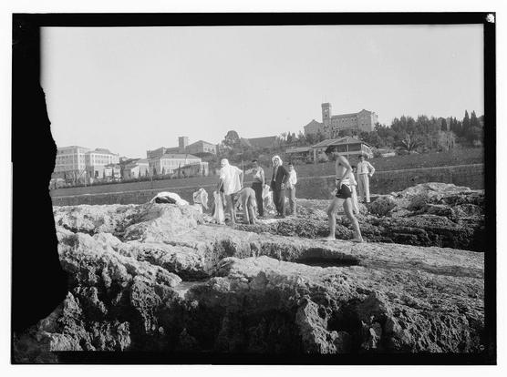 The image is a black and white photograph depicting several individuals on rocky terrain, with the focus seemingly being on leisure or exercise. There are at least eight people visible in various poses: some standing still while others appear to be walking or climbing among the rocks.

In the background, there's an urban landscape featuring multiple buildings of different architectural styles, including what appears to be a large institutional structure with a clock tower and possibly residential houses. The environment suggests this could be near the coast given the rocky foreground which resembles coastal erosion areas often visited for recreational activities such as walking or running.

The photograph has a vignette effect around its edges, drawing attention towards the center where most of the action is occurring. There's also notable wear at one edge and some dirt on another part of the negative, indicating it may be quite old. The attire of the individuals suggests this photo could date back to early 20th century or later.

The photograph carries with it historical context as indicated by a source reference from American University in Beirut which spans approximately between 1925 and 1946, hinting at its possible origin during that period.