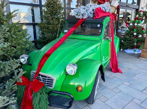 A classic lime green Citroën car has a red ribbon wrapped around it with a flocked Christmas tree and packages on top of it. It's parked in front of the SLO Ranch Marketplace in San Luis Obispo, California.
