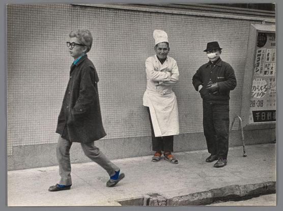 The image depicts three individuals standing on a sidewalk in front of a grid-like background, which may be the exterior wall or facade. On the left is a young man with glasses and distinctive blue shoes walking by; he's dressed casually in a dark coat over a lighter shirt and trousers.

In the middle stands an elderly chef wearing traditional white cooking attire, including a large hat (toque), suggesting he could have been working at a nearby restaurant or kitchen. His expression is relaxed as if observing something with mild interest.

On the right side of the frame is another older man dressed in dark clothing with protective knee-high boots and what appears to be medical crutches; this suggests he may require assistance walking, indicating possible mobility issues like an injury or age-related challenges.

In front of them are signs that include Japanese characters, hinting at a location within Japan. One sign mentions food prices (100 yen), possibly for street vending carts typical in urban areas to feed the working class and tourists alike. The overall atmosphere is casual yet carries a sense of everyday life captured candidly on Dōtonbori Street in Osaka during early 60s, indicative of Ed van der Elsken's style.