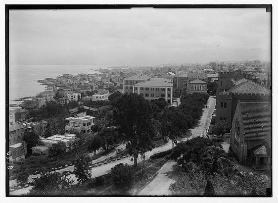 The image is a black-and-white photograph depicting a panoramic view of a cityscape. The scene appears to be from a historical period, judging by the architectural styles and the quality of the photograph. The image shows a mix of residential and possibly institutional buildings with varying roof designs, some with flat roofs and others with pitched roofs. The buildings are closely packed together, and there are trees interspersed throughout the area, providing a sense of greenery amidst the urban environment.
In the foreground, there is a prominent building with a tower and a sloped roof, which could be a church or a significant public building, given its distinct architectural features. The road in the image is lined with trees and appears to be a main thoroughfare, with a few vehicles visible, indicating that the photo was taken before the widespread use of automobiles.
In the background, the city extends towards the sea, which is visible on the left side of the image. The coastline is dotted with more buildings, and further in the distance, there are hills or mountains, adding depth to the landscape. The sky is overcast, with a cloudy appearance, which gives the image a somewhat somber tone. The overall composition suggests a bustling yet orderly urban setting, possibly in the early 20th century.