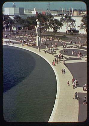 The image depicts an aerial view of a waterfront area with various people walking or sitting along the edge. A prominent statue stands tall on a pedestal, overlooking the water. The surroundings include benches and pathways for pedestrians to enjoy the scenic location.
There are industrial buildings visible in the background behind trees lining the far side of what seems like a river or large lake. This scene appears to be from an event such as World's Fair 1939-40, given its resemblance to architectural elements found at that time and place – specifically references to "Trylon" and "Perisphere," iconic structures related to this fair.
This particular image is credited to Gottscho-Schleisner, Inc., a photography company known for documenting various events throughout the 20th century. The photo measures approximately two inches by two inches in size with color print quality on thin film stock.
