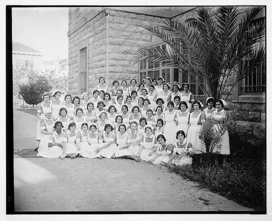 The image depicts a large group of women, presumably nurses, from an early 20th-century era. They are dressed in nurse uniforms consisting of white dresses with aprons and head coverings that resemble traditional nursing caps or bonnets. The attire suggests the photograph is historical, as it reflects the typical uniform style for nurses during this period.

The group is arranged outdoors on a paved surface adjacent to what appears to be an institutional building made of stone. Behind them stands a large palm tree with long fronds extending toward the camera's right side. To their left are more trees and shrubbery, indicating they may be in a courtyard or garden setting associated with the institution.

The women sit on the ground, some kneeling, while others stand to form rows behind those seated. Their expressions range from neutral to smiling as they pose for what seems like an official group photo session. The photograph is black-and-white and has a grainy texture typical of early photography technology.

There are no visible texts or inscriptions within the image that provide further context about these individuals, their institution, or location at the time it was taken.