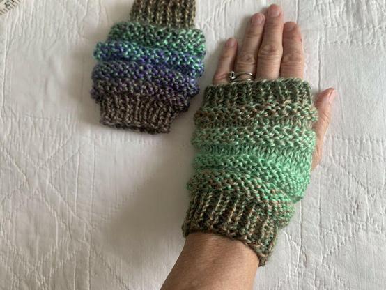 Photo of a left hand wearing green and blue knit fingerless gloves. The other glove lays flat on the left.