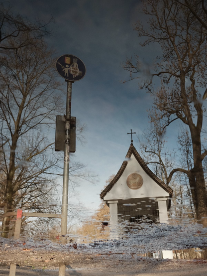 A picture of a street sign next to a small chapel under a cloud-spotted sky. Empty winter trees in the background.
The image seems rather pixelated at the bottom, the top is slightly distorted almost like in a dream.
Once you turn the image up side down, you realize, it’s a mirrored image taken against a puddle.