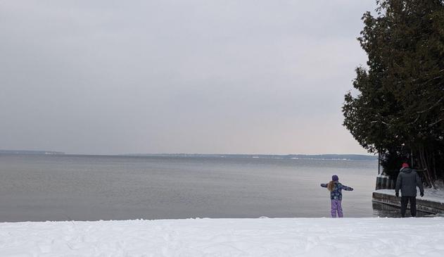 A snow-covered beach on Lake Simcoe with the cold grey water just beyond the white shoreline.  Granddaughter and her Dad, bundled up against the cold, are in the far right near a concrete pier.