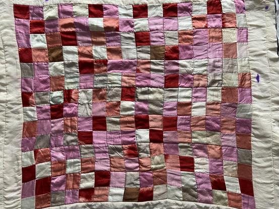A small quilt of two-inch squares in a semi-random distribution of dark red, pink, orange, and pale sage green. It has a cream colored border with a purple stain. It has a lovely sheen because it's pure silk.