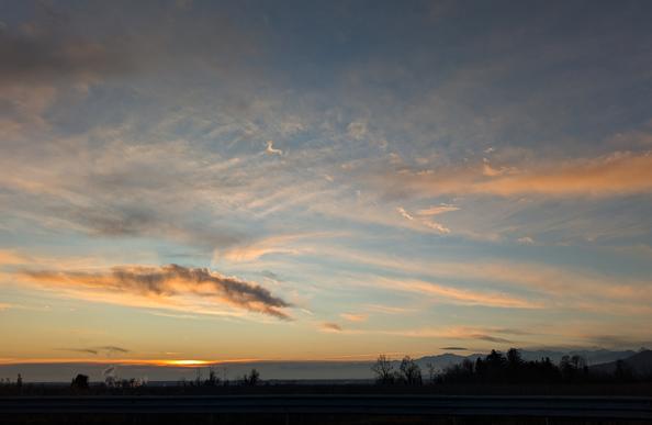L'immagine mostra un tramonto panoramico caratterizzato da un cielo spettacolare e colorato su un paesaggio che include campi, una linea di alberi e montagne sullo sfondo.
Il cielo presenta una ricca gamma di tonalità, passando da un blu profondo nella parte superiore a strisce vibranti di arancione, giallo e rosa vicino all'orizzonte. Questo fenomeno si verifica perché, al tramonto, la luce solare attraversa uno strato più spesso di atmosfera, disperdendo le lunghezze d'onda più corte (blu e verde) e lasciando prevalere i colori caldi.
Sono visibili vari tipi di nuvole: quelle più alte e sottili (probabilmente cirri) sono illuminate dalla luce del sole, mentre una formazione nuvolosa più densa e scura si trova più in basso a sinistra, apparendo scura perché in ombra rispetto alla posizione del sole.
Il primo piano scuro suggerisce la sagoma di un guardrail stradale e terreni aperti. Una linea di alberi e, più in là, la silhouette di una catena montuosa completano la composizione, offrendo un contrasto netto contro il cielo luminoso