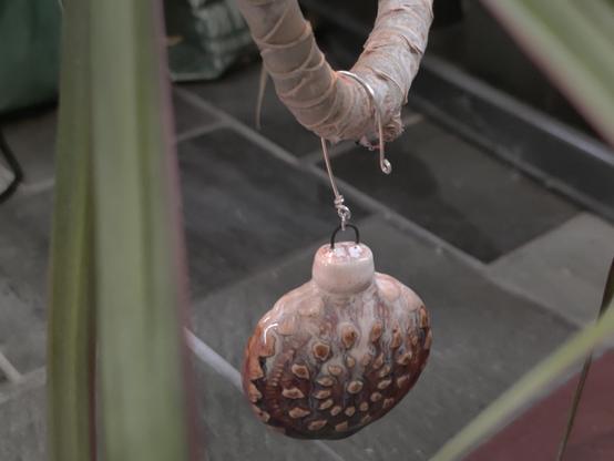 Looking through some long foreground leaves at a crook of a houseplant's trunk, where a holiday tree ornament is hanging. The ornament is round with intricate doily-like patterns in the ceramic face. The glaze is a swirl of beautiful white, purple, red, and orange. It's hanging from a silver looped hook.