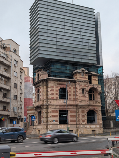 An old building that looks like it was built around the 1900s, large stones in sandstone and brick-red colouring, sits on a street corner in a city. The natively two-storey building speaks of long gone prosperity, however above it several more storeys throne above it in modern steel and glass fashion. The add-on is slightly taller than the original building structure