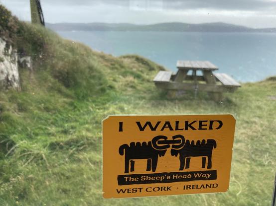 Durch eine Glasscheibe blickt man auf Gras und eine Picknickbank-Tisch-Kombination hoch über einer blauen Bucht. Auf der anderen Seite Hügelkette einer Halbinsel. Auf der Glasscheibe der gelbe Aufkleber: I WALKED THE SHEEP'S HEAD WAY, WEST CORK - IRELAND sowie das Logo mit zwei Schafsböcken, die einander gegenüber stehen,