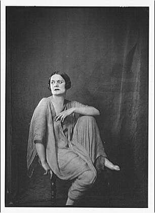 This black and white photograph features a woman seated on a stool with her legs crossed at the ankles, dressed in loose-fitting clothing. She appears contemplative or poised for an artistic portrayal, as she gazes off to the side rather than directly into the camera lens.
The image is credited to Arnold Genthe (1869-1942), and it's labeled "Irma Duncan dancer portrait photograph 1929 Jan. 29." It was taken with a single negative on safety film, measuring 5 x 7 inches in size.
A more detailed description of the woman includes her sitting position on what seems to be a round stool or chair, with one arm resting along it and the other extended outwards as she looks away from the camera. Her attire suggests an informal yet stylish approach, consisting of loose-fitting trousers paired with a draped shawl-like garment over her shoulders.
The overall composition is simple but evocative, capturing a sense of elegance and introspection through its monochrome tones and thoughtful subject placement within the frame.