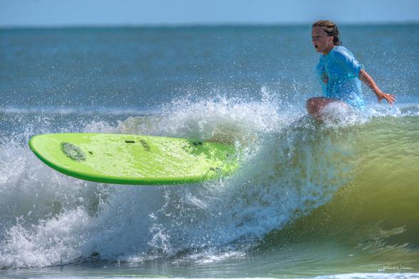 Surfer in bright blue rash guard paddling through wave with green surfboard, backlit by brilliant water and sky with shadow across face.