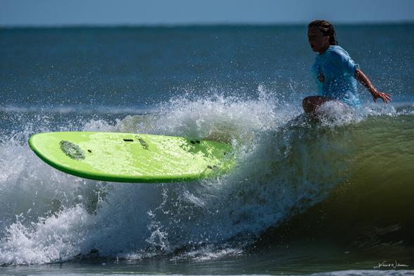 Surfer in bright blue rash guard paddling through wave with green surfboard, backlit by brilliant water and sky with shadow across face.