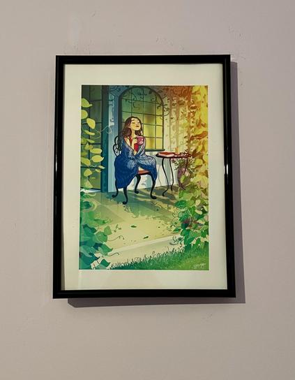 A framed print shows a young woman sitting on the patio outside her house on a sunny day, wearing pyjamas and wrapped in a blanket, lifting her face (eyes closed) to soak in the sun and holding a big mug of tea or coffee in her hands. Lush green plants from the garden frame the patio and leave leafy shadows on her house.
