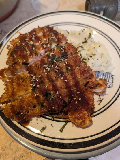 A fried chicken cutlet resting on a mound of mashed cauliflower. The cutlet has tonkatsu sauce over top and furikake sprinkled over everything.