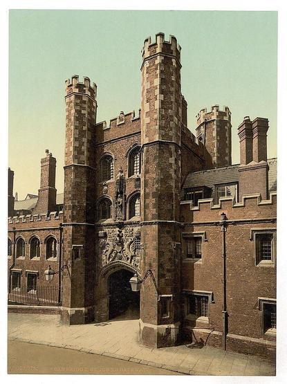 The image shows a historic building with two prominent, cylindrical towers capped with battlements. These towers flank an arched entrance featuring detailed stone carvings and statues, suggesting a medieval or gothic architectural influence. The structure is made of brickwork with some sections displaying yellowish hues possibly due to the weathering over time. Several rectangular windows are visible on both levels of the building's facade, including smaller square openings near the top.

In front of the entrance stands a street lamp and what appears to be part of a gated courtyard or garden area enclosed by low walls, separated from the sidewalk by railings. The sky is pale blue with no significant cloud cover, indicating clear weather conditions at the time the photograph was taken.

The setting seems urban based on the paved surface alongside the building's exterior wall. A faint outline suggests additional buildings adjacent to this historic structure. Text overlaid at the bottom of the image indicates "Cambridge St. Johns Gate," identifying the location as a specific entryway in Cambridge, England, associated with an educational institution known for its historical and architectural significance.

The overall condition appears well-preserved yet aged, suggesting that while it has been maintained over time, the building retains features characteristic of its era, including robust construction materials and design elements typical of older English architecture.