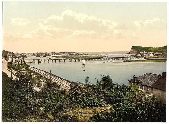 The image depicts a tranquil coastal scene, likely captured in late 19th or early 20th-century Britain. In the foreground, there's lush greenery with small structures that resemble residential houses nestled among trees and shrubs. A railway track is visible on the left side of the photo, hinting at an active transportation route during this era.

The middle ground showcases a vast expanse of water extending to the horizon where it meets the sky. The calm sea has several boats dotting its surface, suggesting maritime activity typical for coastal areas in that period. A prominent feature is a long pier or jetty stretching into the ocean on the right side, with structures at its end and supports visible beneath.

To the left of this structure stands another bridge spanning across the waterway towards what appears to be an industrial port area filled with ships docked along piers. The backdrop features rolling green hills under a partly cloudy sky. A notable hill rises in the distance on the right, adding depth to the landscape.

The overall tone and coloration suggest that this is a photograph or postcard printed using photogravure technique from an archive collection titled "Shaldon Bridge," indicating its location as Teignmouth, Devon, England. The image captures both everyday life by the sea with industrial elements and natural beauty of coastal scenery during a specific historical period in Britain.