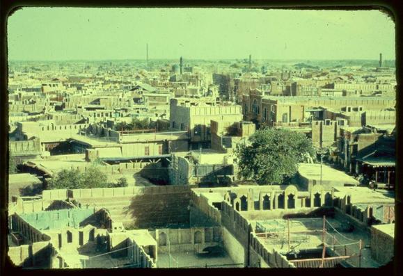 The image depicts an aerial view of a densely built urban area with numerous buildings, many featuring traditional architectural elements. The structures vary in design and size but predominantly consist of rectangular forms with flat roofs or sloped terraces. Some buildings have prominent arched doorways and windows, indicative of historical Islamic architecture.

In the foreground, there's an open courtyard enclosed by low walls, which could be part of a larger complex like a mosque or madrasa (educational institution). The area is populated with smaller courtyards surrounded by higher walls. A tree stands out among these structures, providing contrast to the built environment.

The middle ground shows more buildings and streets, creating a layered urban landscape that extends towards the horizon where modern industrial elements such as chimneys or towers can be seen against an expansive sky. The color palette is warm with varying shades of brown and beige dominating the scene, suggesting either the time of day—likely midday due to the brightness—or the weather conditions.

There's no visible activity on the streets from this vantage point, which could imply a quiet moment or that the photo was taken during non-peak hours. The overall impression is one of an ancient city with a rich architectural heritage amidst modern development.