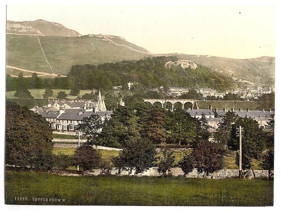 The image depicts a picturesque view of a town nestled in a valley, with a backdrop of rolling hills and a prominent mountain range. The town appears to be a quaint settlement with a mix of residential and possibly commercial buildings, featuring pitched roofs and chimneys. There are trees scattered throughout the area, adding greenery to the scene, and a stone wall runs along the foreground, separating the viewer's perspective from the town.

In the mid-ground, a railway viaduct with multiple arches spans across the scene, suggesting the presence of a railway line that connects the town. The architecture of the buildings is traditional, with some structures having a church-like appearance, indicated by a spire visible in the distance. The overall atmosphere is serene and rural, with a sense of historical charm suggested by the style of the buildings and the vintage feel of the image. The lighting and coloration give the impression of a photograph taken in the late 19th or early 20th century.