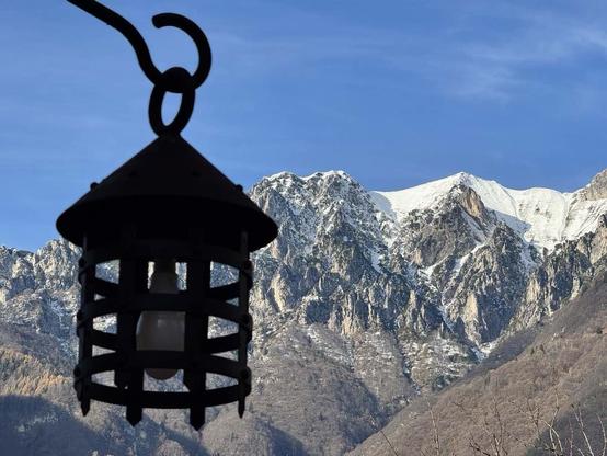 A wrought iron lantern in the foreground with a mountainous landscape in the background, featuring snow-capped peaks under a clear blue sky.