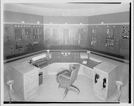 The image depicts an interior view of a control room or a similar operational setting, likely from a mid-20th century facility. The room is dominated by a large, curved desk in the center, which appears to be the primary workstation. On this desk, there are two typewriters, one on each side, suggesting a setup for communication or record-keeping. The desk is surrounded by a high-backed, swivel chair, positioned in front of the desk.
The walls are lined with numerous control panels, each equipped with various switches, dials, and indicators. These panels are arranged in rows and columns, indicating a highly organized and systematic approach to managing operations. The design of the panels suggests they are used for monitoring and controlling mechanical or electrical systems, possibly related to power generation or industrial processes.
The overall lighting is even, and the room appears clean and well-maintained. The color palette is muted, with shades of gray and beige, typical of the industrial design of the time. The image has a vintage quality, consistent with photographs from the mid-20th century. The photograph itself has a black-and-white tone, adding to the historical feel of the scene.