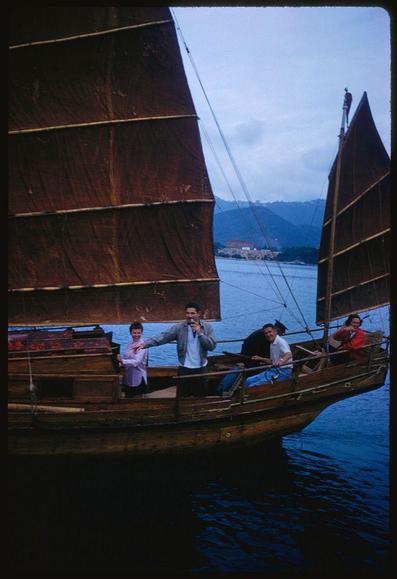 The image shows a group of people on a traditional Chinese junk boat. The boat is characterized by its large, dark brown sails, which are partially unfurled, indicating that it is either in motion or preparing to move. The sails have a distinct layered structure, with horizontal folds. The boat itself is wooden, with a traditional design, and has a flat bottom. The people on the boat are dressed in light clothing, suggesting a casual outing or tour. One person is holding a microphone, possibly indicating that they are giving a speech or leading a tour. The background features a body of water, with a distant shoreline and hills covered in greenery. The sky is overcast, giving the scene a soft, diffused light. The overall setting suggests a serene and picturesque environment, likely a tourist attraction.