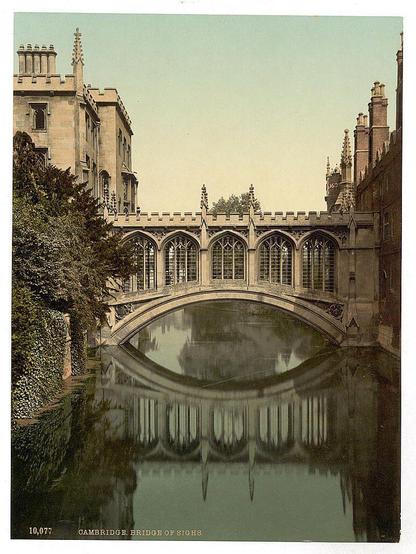 The image displays a picturesque view of an ornate bridge over water, reflecting its architecture on the surface below. The structure is medieval in style with pointed arches and large windows that hint at Gothic influences commonly found in European historical buildings, particularly those associated with universities or old institutions. Surrounding the bridge are two imposing stone structures adorned with intricate stonework, including pinnacles that suggest a fortified design typical of collegiate architecture. Ivy partially covers one side of these edifices, adding to their ancient charm. The water beneath mirrors almost perfectly the silhouette and details of both the bridge and surrounding buildings. Above this scene is a clear sky in soft hues, possibly indicating either dawn or dusk due to its gentle coloration. This image seems like an old postcard from Cambridge University's Bridge of Sighs, capturing not just the architecture but also evoking a sense of history and academia associated with such settings.