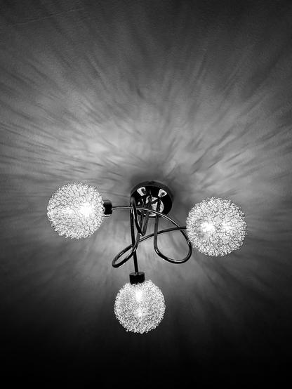 Black and white image of a modern, twisting ceiling light featuring three fine metallic globes on the end of smooth, curving and intertwining stems which are casting intricate light and shadow patterns on the ceiling
