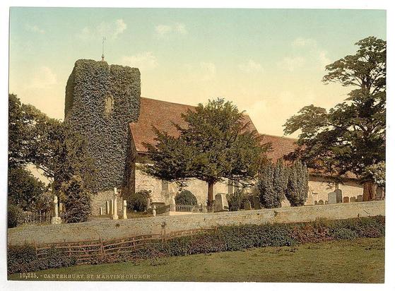 The image depicts an old church with a distinctive feature: its tower is almost completely covered in ivy, giving it a rustic and ancient appearance. The building has a stone structure with visible windows framed by the climbing foliage. Adjacent to the main body of the church, there's a smaller section that could be part of additional structures or extensions. 

A red-tiled roof extends from one side of the tower to what appears to be an adjoining nave or sanctuary. Surrounding the church is a fenced-off graveyard with numerous gravestones and headstones scattered throughout, suggesting it has been in use for some time.

The foreground features neatly trimmed bushes and possibly a garden area, enclosed by a simple fence that separates the grounds of the church from what could be another property or public space. The background shows trees under a sky transitioning into dusk, with warm colors indicating either sunrise or sunset light casting an orange hue across the scene.

This image likely comes from around the late 19th to early 20th century based on its style and historical context as indicated by "Views of the British Isles England Cambridge" and provided date range.