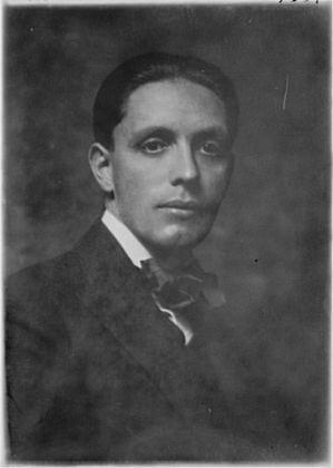The image is a black-and-white portrait of a man. The photograph appears to be vintage, likely from the early to mid-20th century, given its style and the quality of the image. The man is dressed formally, wearing a dark suit with a white shirt and a bow tie. His hair is neatly combed, and he has a serious expression on his face, looking directly at the camera. The background is plain and dark, which helps to emphasize the subject. The lighting is focused on the man's face, creating a slight shadow on the right side of his face. The overall tone of the image is formal and dignified.
