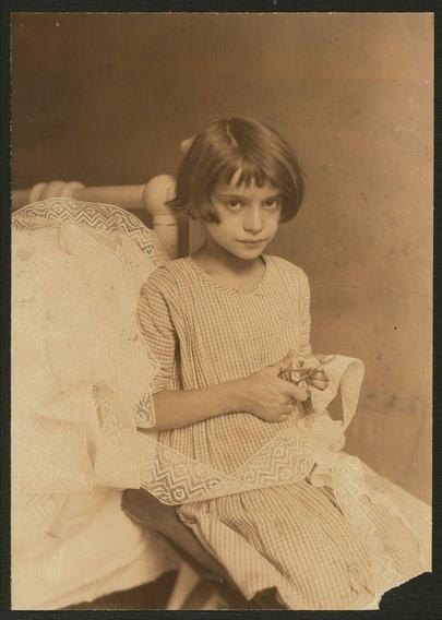 The image appears to be a sepia-toned photograph, likely from the early 20th century, given its style and the quality of the photograph. It features a young girl seated in a chair, wearing a dress with a checkered pattern. The dress is light-colored, possibly beige or cream, with a delicate white lace trim at the bottom, which extends over the edge of the chair. The girl has shoulder-length hair with bangs, and her expression is neutral or slightly serious. She is holding a pair of scissors in her hands, which are positioned near a piece of fabric or lace in her lap. The background is plain and dark, which helps to focus attention on the subject. The overall tone of the image is nostalgic, evoking a sense of a bygone era.