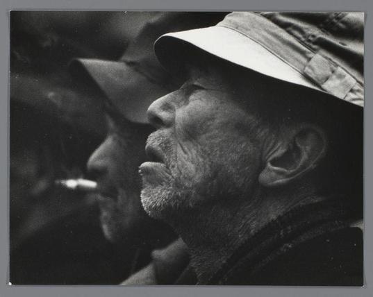 This image is a black and white photograph featuring a close-up of a man's profile. The man appears to be wearing a wide-brimmed hat, which casts a shadow over his face. His facial features, such as his nose, mouth, and ear, are prominently visible. The man has a beard and mustache, and his skin shows signs of age with visible lines and texture. The background is blurred, suggesting that the focus is on the man's face. The lighting is dramatic, with strong contrasts between light and shadow, giving the photograph a moody and intense feel. The overall composition and style of the image suggest it could be a portrait or a candid shot.