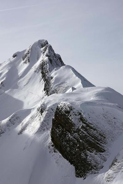 Ein schneebedeckter, steiler, felsiger Gipfel dominiert das Bild, die Felsen schauen teils dunkel aus dem weißen Schnee heraus.
