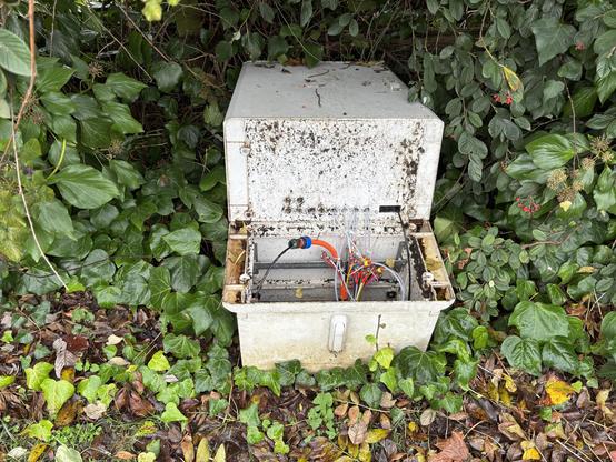 A roadside telecoms connection box that’s been broken open (maybe a car drove into it) the top half appears to be connected with hinges to the bottom half and there are many wires/fibres between the two