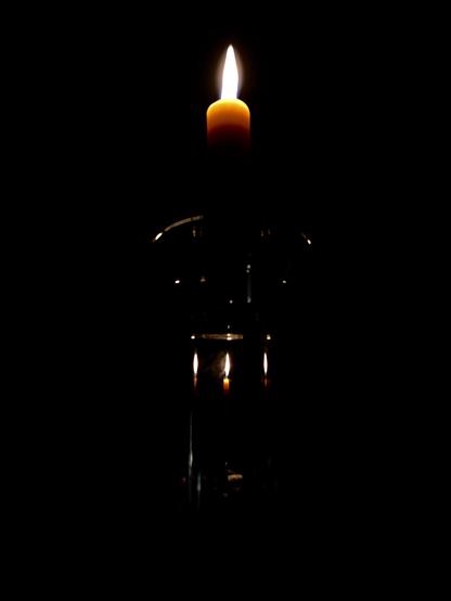 Photo of a lit candle in a glass candlestick against a completely black background. The small flame points directly upward and casts a soft glow on the top of the gold-brown wax. Light glints at the edges of the glass holder, and through its main column we can see three refracted images of a second lit candle, which sits further from the camera and directly behind the holder. The only illumination comes from the flames of the candles.